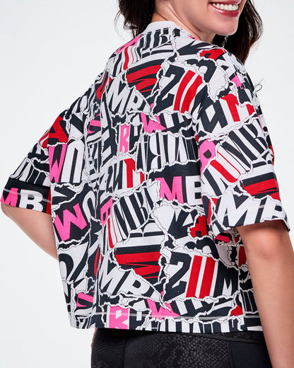 A close-up back view of a female model wearing a short-sleeve crop top with a bold abstract ZUMBA text pattern print in black, white, red, and pink.