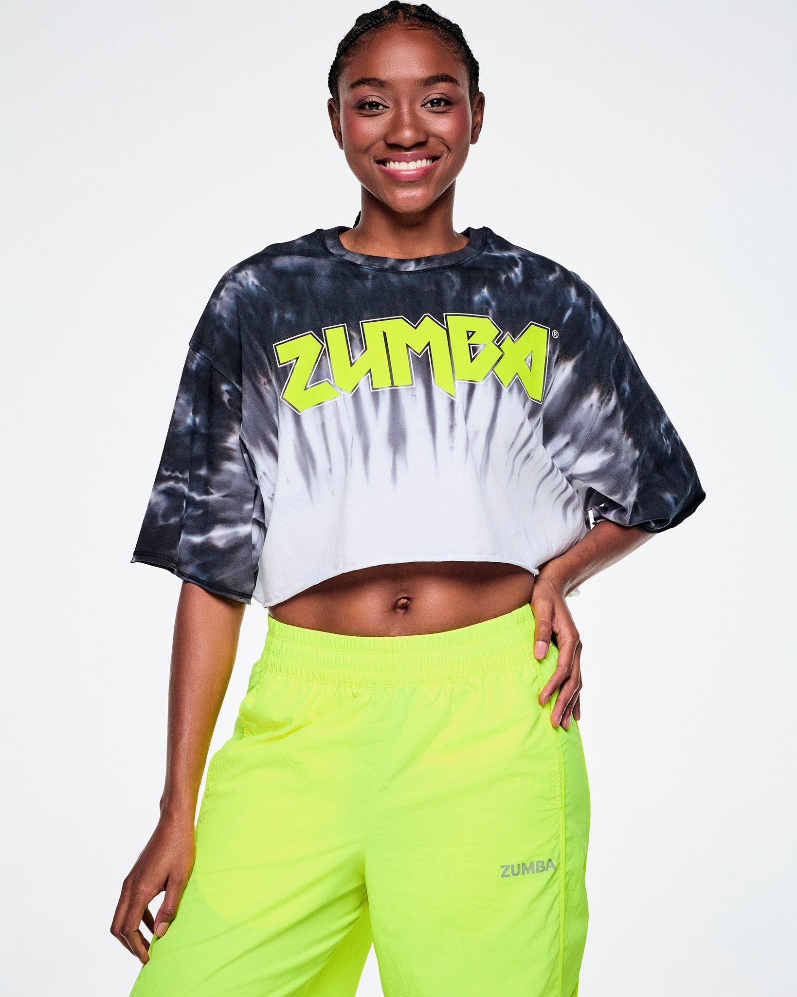 A female model wearing a cropped tie-dye Zumba top with neon yellow 'ZUMBA' rock-like text across the chest, paired with yellow pants.