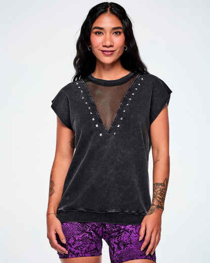 A female model wearing a acid-washed patterned black raw edge short-sleeve top with a mesh V-neckline and silver studs, paired with purple snake-skin patterned biker shorts, against white background.