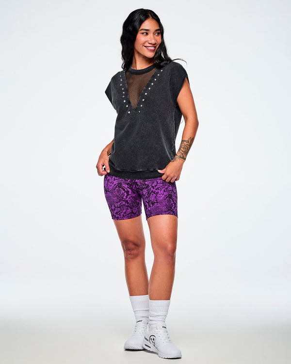 A female model wearing a acid-washed patterned black raw edge short-sleeve top with a mesh V-neckline and silver studs, paired with purple snake-skin patterned biker shorts and white shoes and socks.