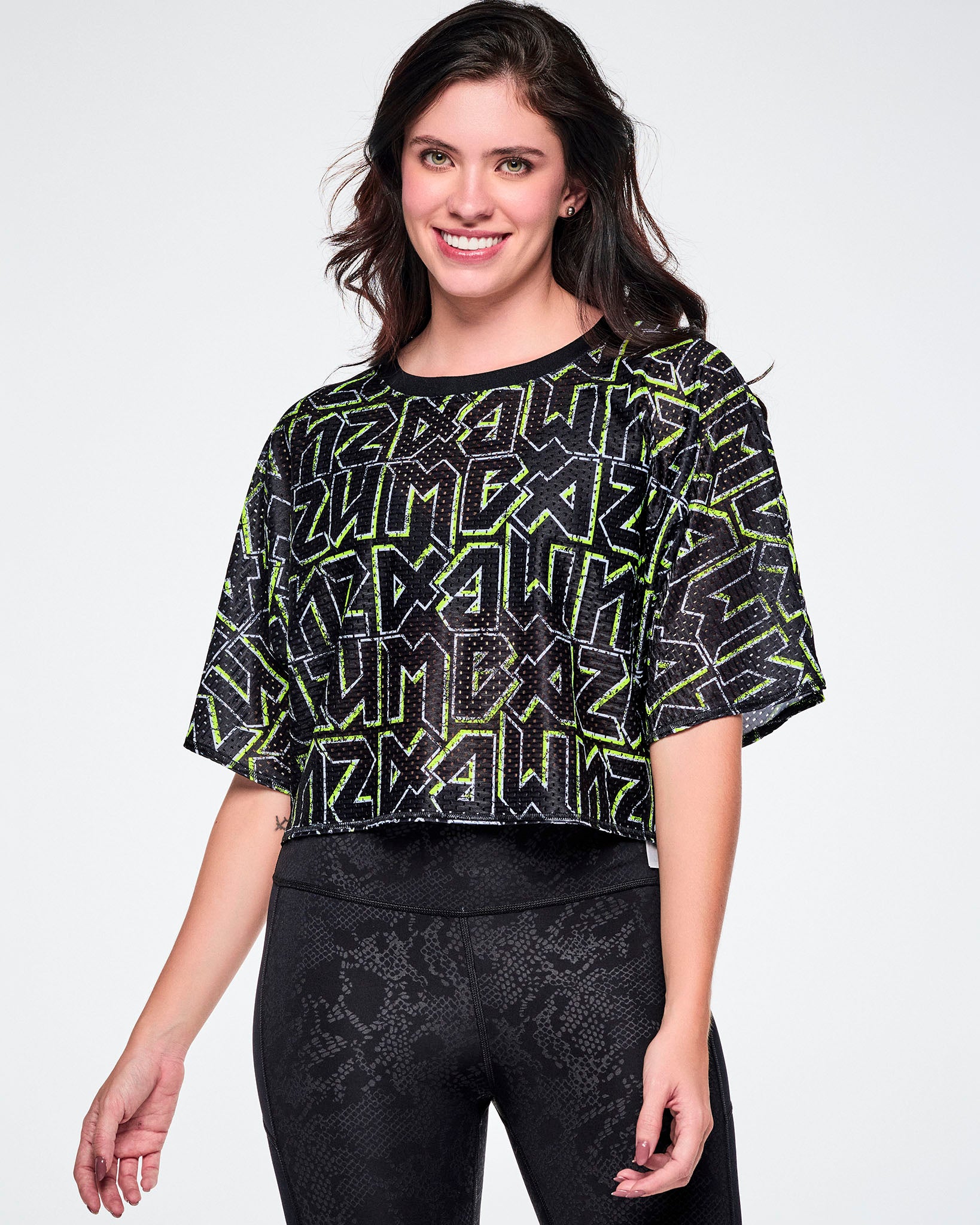 A female model wearing a short-sleeve mesh boxy crop top with rock-like letter patterning that says "ZUMBA" in black and outlined in green, and paired with black leggings.