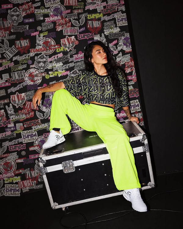 A female model wearing neon yellow-green pants, a matching black and green mesh top and white shoes, as she sits on a black equipment case in front of a wall covered in Zumba rock emblems.