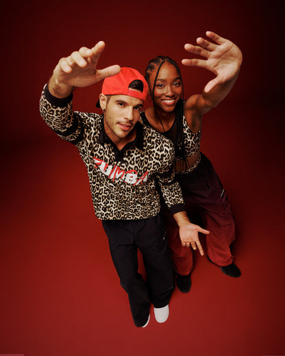 A male and female model posing against a solid dark red studio background. The male model on the left wears a leopard-print long-sleeve shirt with 'ZUMBA' printed in red and white across the chest, black pants, and a red cap. The female model on the right wears a tan leopard print bra and gradient red and black pants.