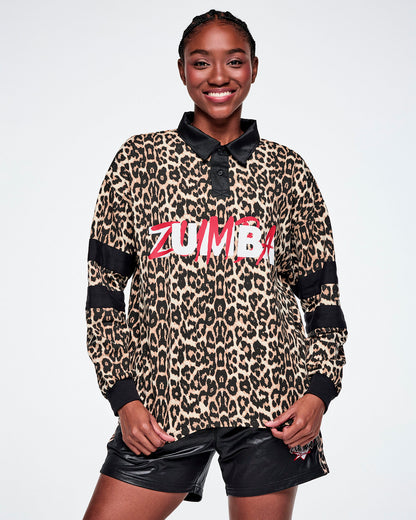 A female model wearing a long-sleeve leopard-print polo shirt with black collar, cuffs, and sleeve stripes, featuring red and white 'ZUMBA' text across the chest, paired with black shorts against a white background.
