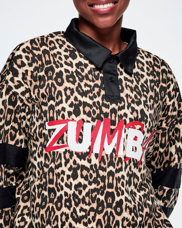 A close-up of a female model wearing a Zumba long-sleeve polo shirt with a beige and black leopard print, black satin collar and cuffs, and large red and white 'ZUMBA' text on the front.