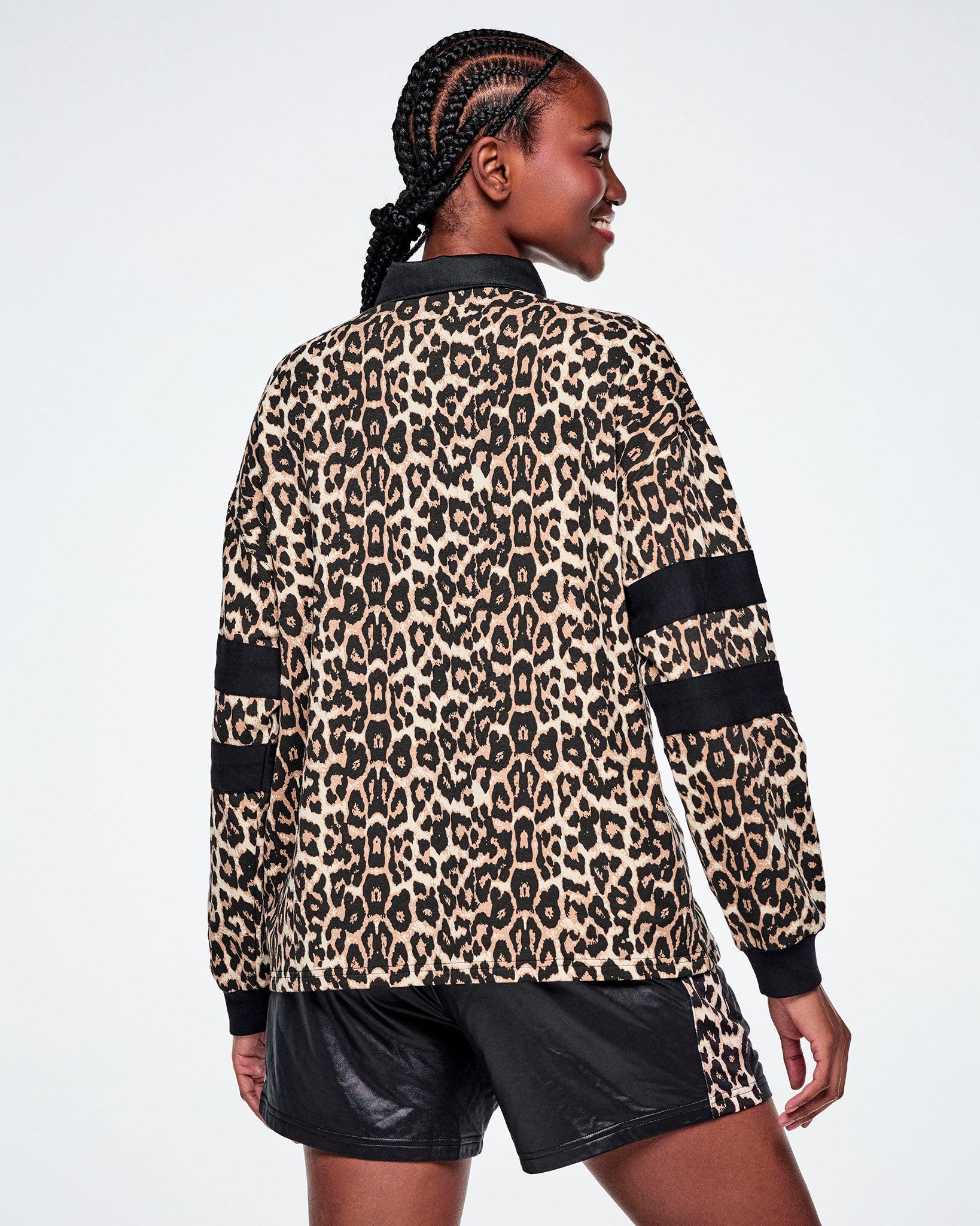 Back view of a female model wearing a long-sleeve leopard-print polo shirt with black collar, cuffs, and sleeve stripes, paired with black shorts against a white background.