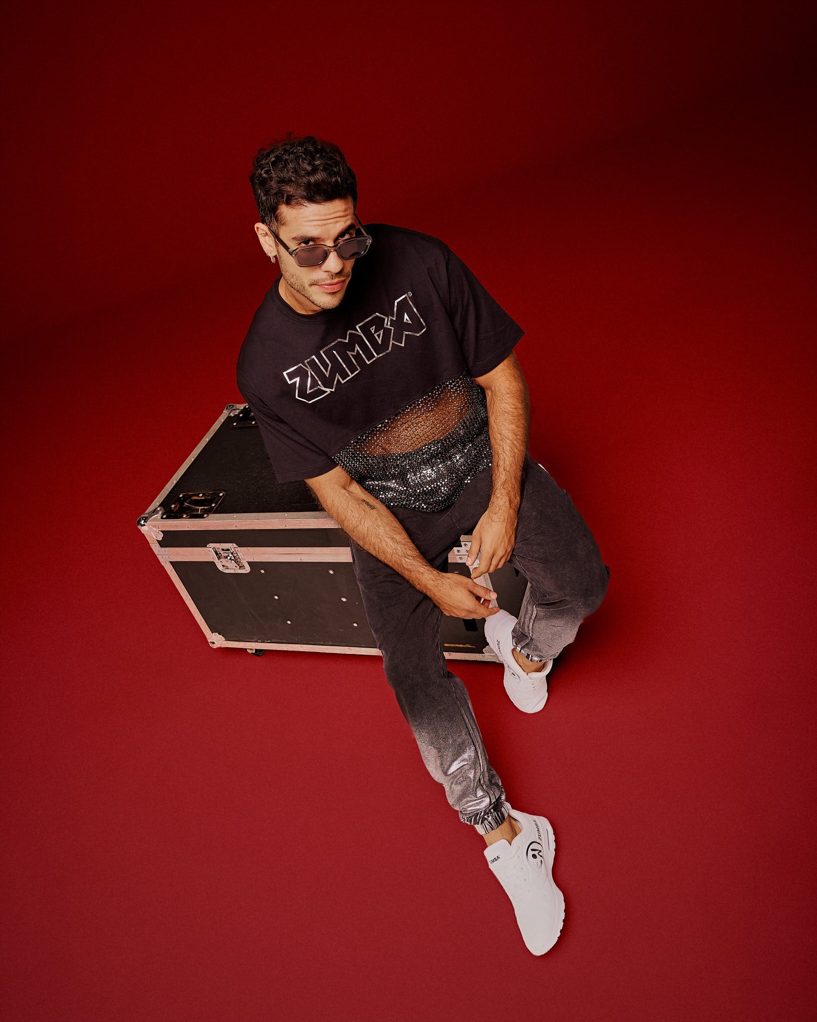 A male model sits on a black equipment case against a solid red studio background, wearing a black Zumba cropped top with a sheer mesh panel with sequins and a large silver outlined ZUMBA logo across the chest, paired with black pants and white shoes.