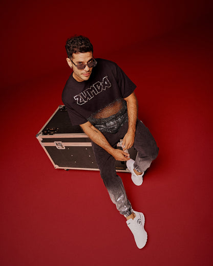 A male model sits on a black equipment case against a solid red studio background, wearing a black Zumba cropped top with a sheer mesh panel with sequins and a large silver outlined ZUMBA logo across the chest, paired with black pants and white shoes.
