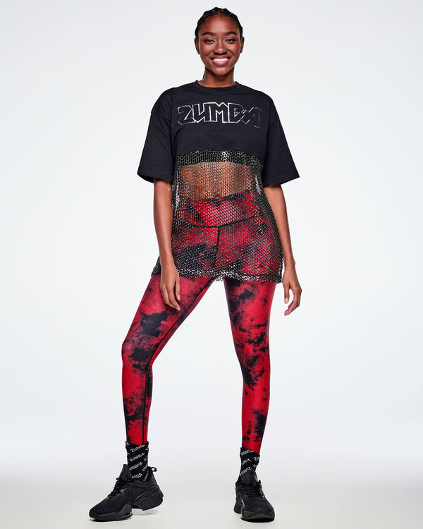 Full view of a female model wearing a black Zumba cropped top with a sheer mesh panel with sequins and a large silver outlined ZUMBA logo across the chest, paired with red and black leggings, black socks and shoes.