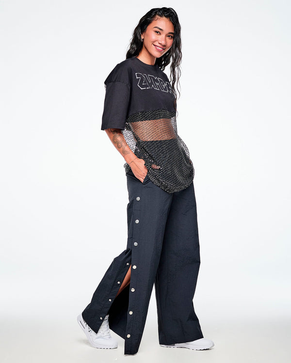 Full view of a female model wearing a black Zumba cropped top with a sheer mesh panel with sequins and a large silver outlined ZUMBA logo across the chest, paired with black pants with side snaps and white shoes.