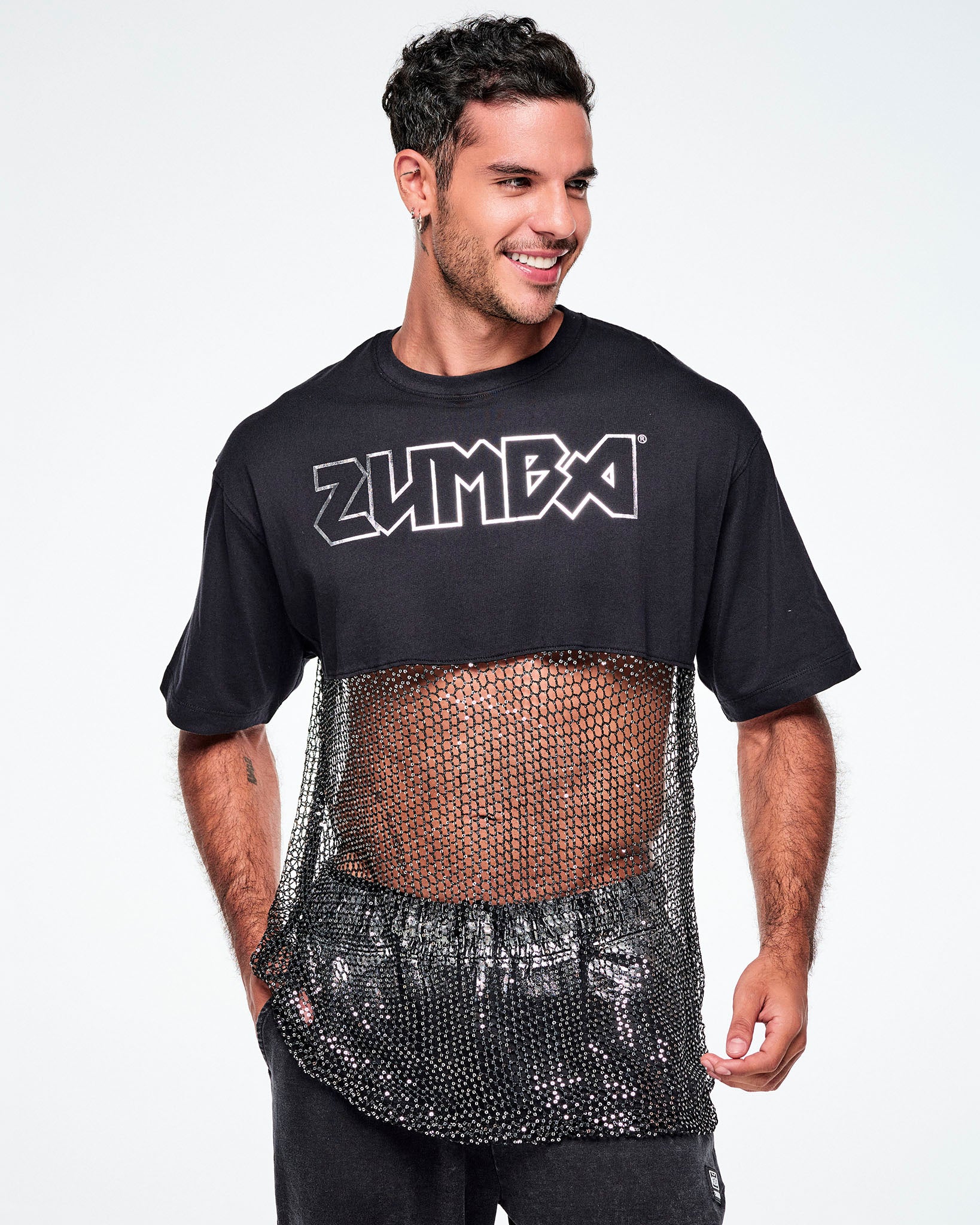 A male model wearing a black Zumba cropped top with a sheer mesh panel with sequins and a large silver outlined ZUMBA logo across the chest, paired with black pants.