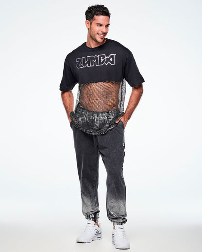 Full view of a male model wearing a black Zumba cropped top with a sheer mesh panel with sequins and a large silver outlined ZUMBA logo across the chest, paired with black pants and white shoes.