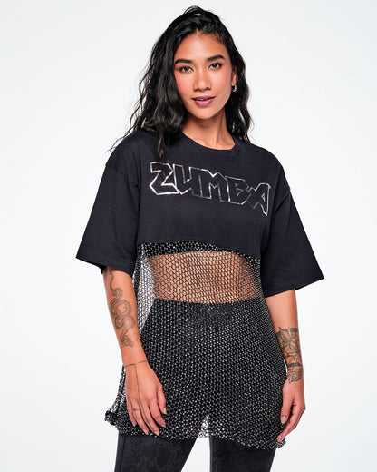 A female model wearing a black Zumba cropped top with a sheer mesh panel with sequins and a large silver outlined ZUMBA logo across the chest, paired with black leggings.