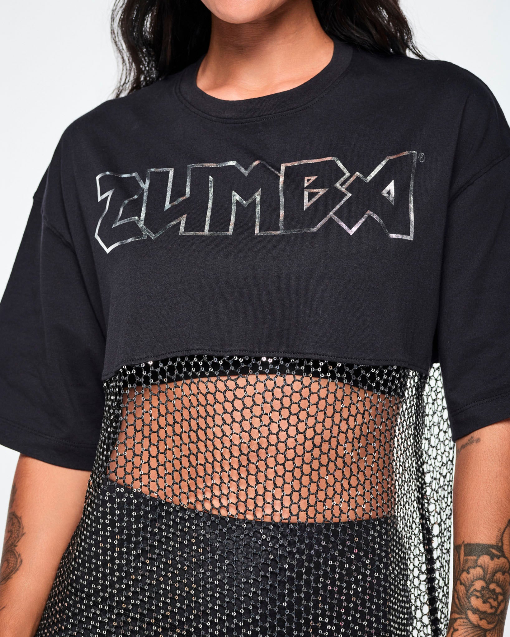 Close-up of a female model wearing a black Zumba top with a sheer mesh panel with sequins and a large silver outlined ZUMBA logo across the chest.