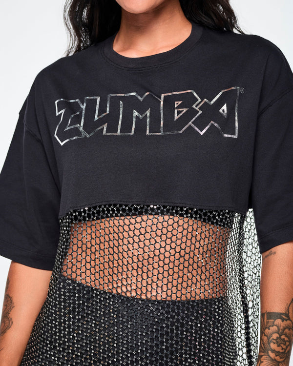 Close-up of a female model wearing a black Zumba top with a sheer mesh panel with sequins and a large silver outlined ZUMBA logo across the chest.