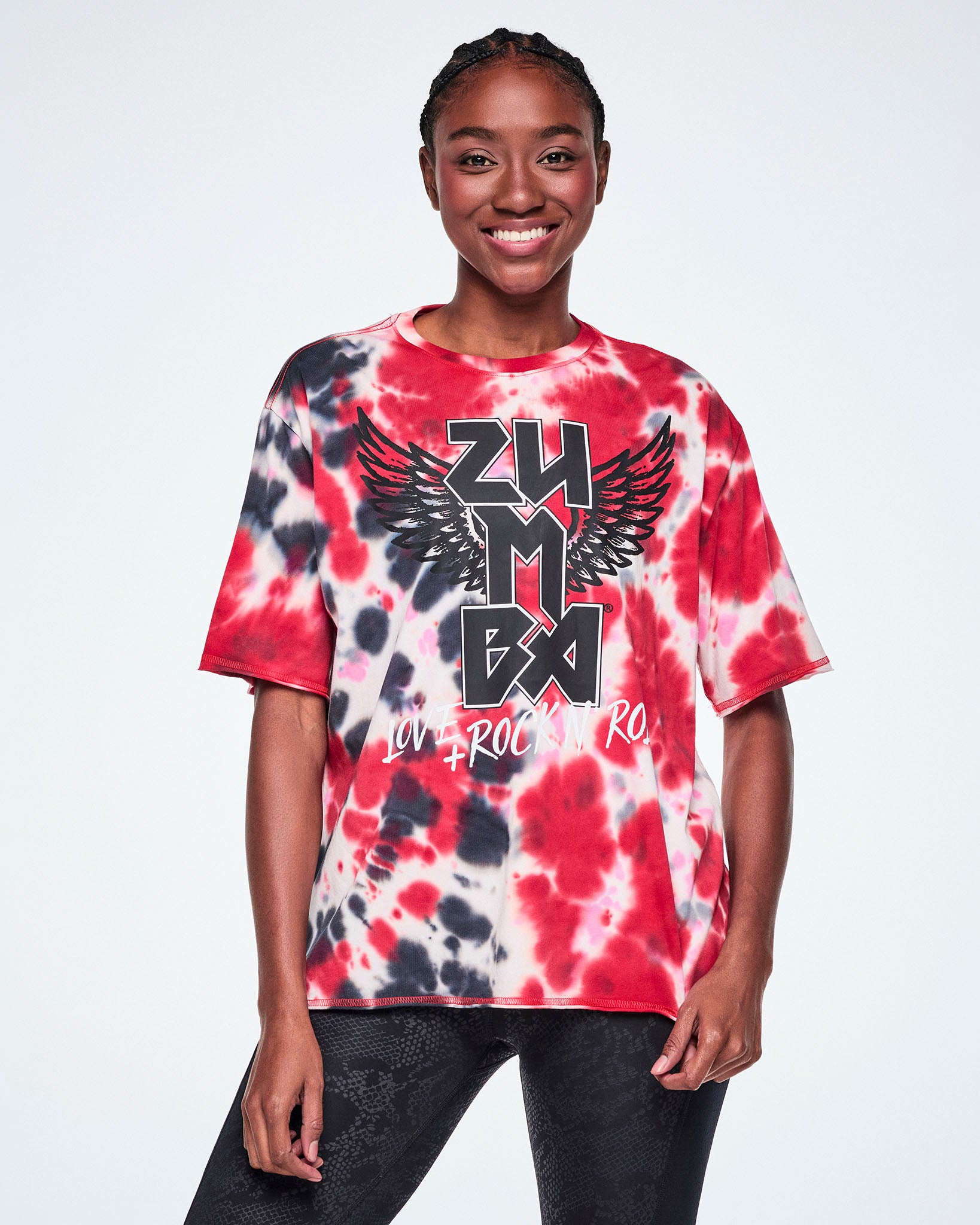 A female model wearing an oversized red, white, and black tie-dye patterned tee with a large black winged 'ZUMBA' rock / metal like logo and white 'LOVE+ROCK N' ROLL," paired with black leggings.