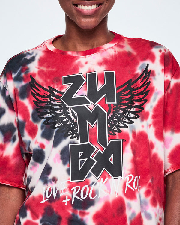 A close-up of a female model wearing a red, black, and white tie-dye tee with a large black and white ZUMBA logo with wings on the front, and the text 'LOVE + ROCK IT!' below.