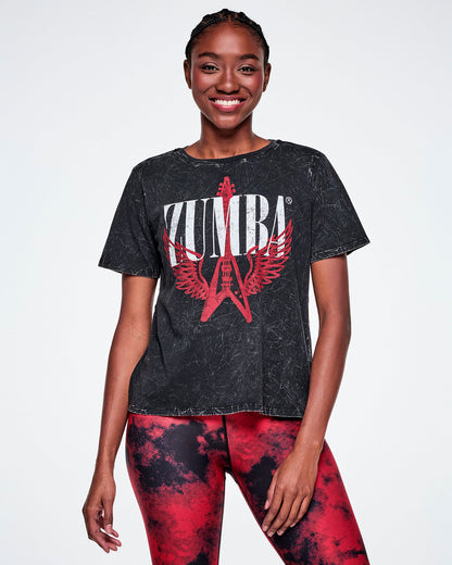 A female model wearing a black acid-washed patterned Zumba graphic tee with a red guitar with wings logo and ZUMBA in big white text, paired with red and black leggings.