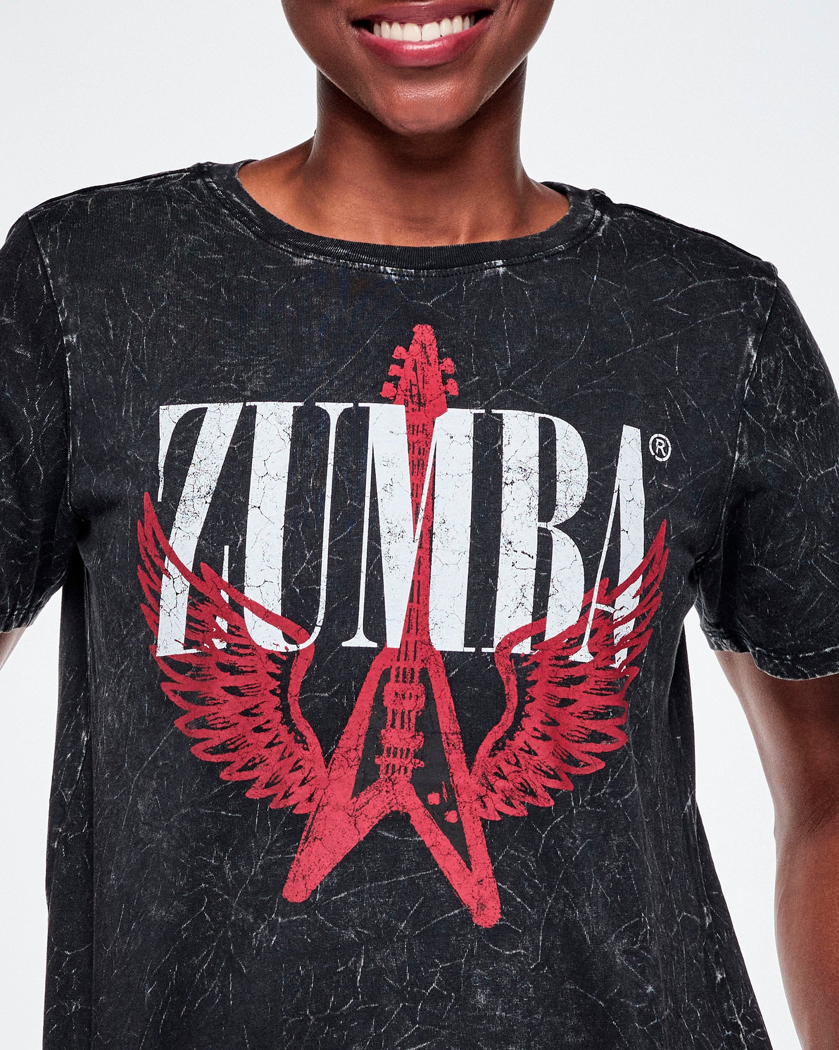 Close-up of a female model wearing a black acid-washed patterned Zumba graphic tee with a red guitar with wings logo and ZUMBA in big white text.