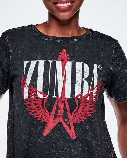 Close-up of a female model wearing a black acid-washed patterned Zumba graphic tee with a red guitar with wings logo and ZUMBA in big white text.