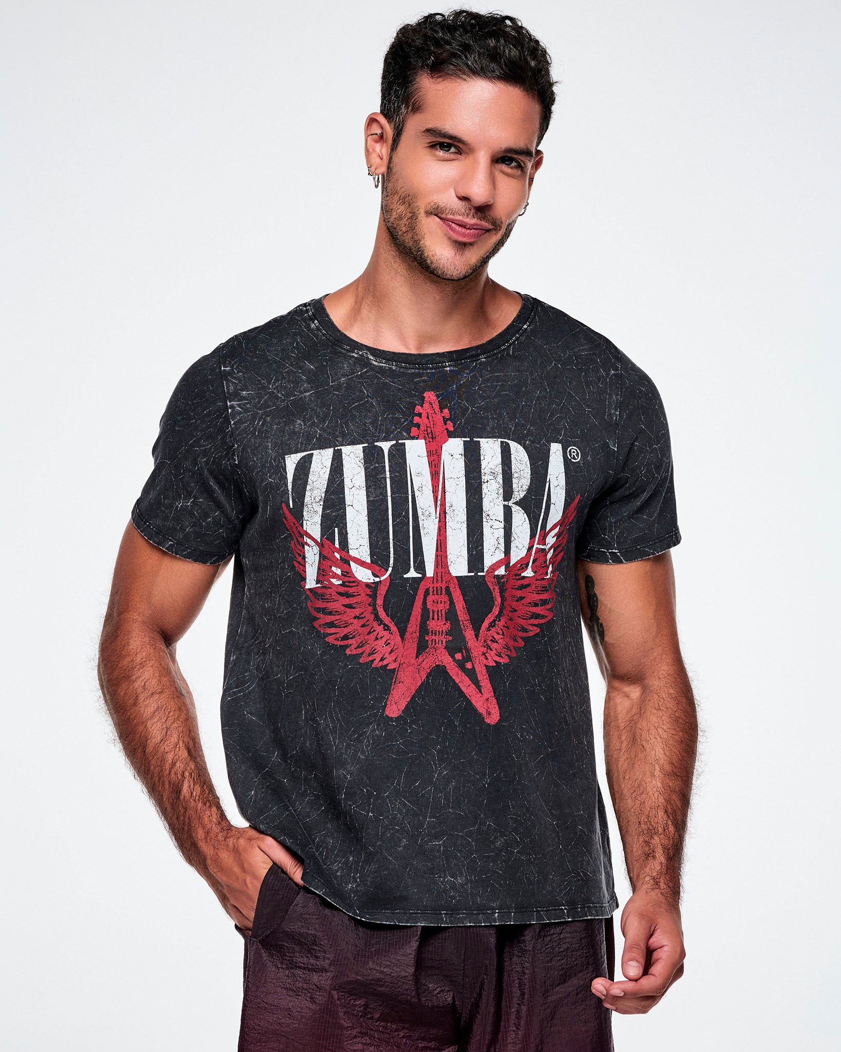 A male model wearing a black acid-washed patterned Zumba graphic tee with a red guitar with wings logo and ZUMBA in big white text, paired with gradient red and black pants.