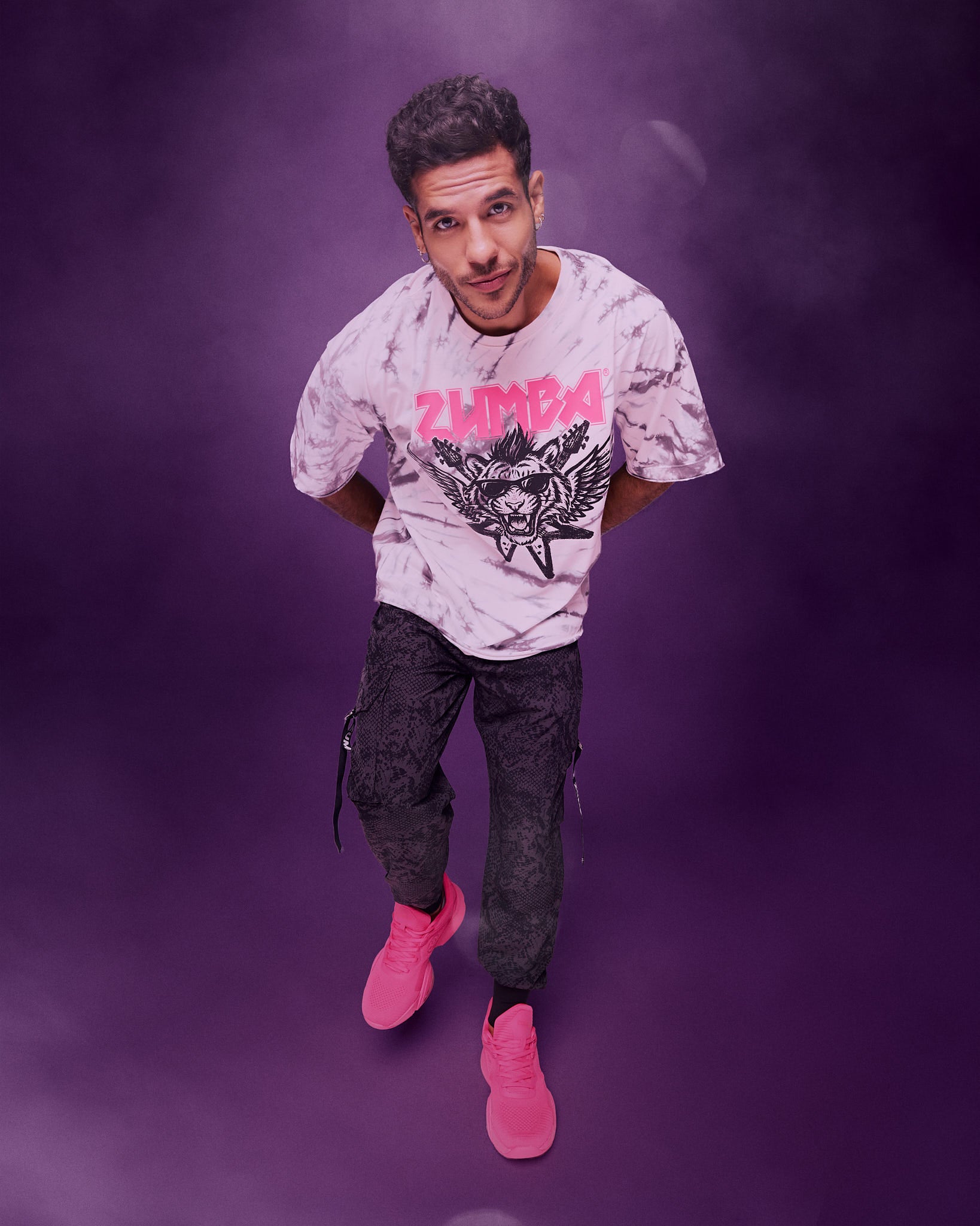 A male model wearing a white and gray tie-dye patterned graphic tee with a tiger in sunglasses and guitars and angel wings behind, paired with black pants and pink shoes, posing against a purple studio background.