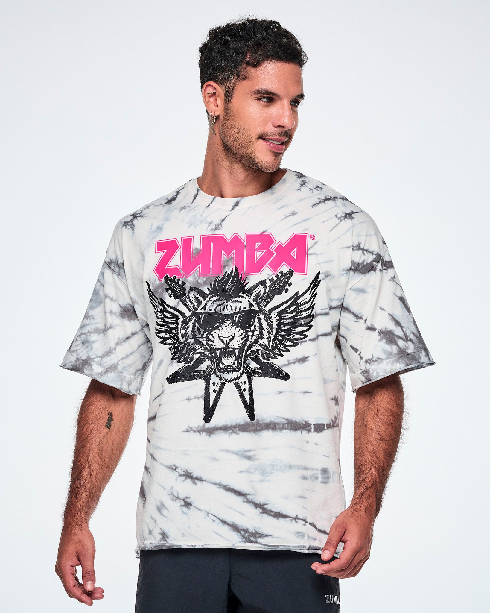 A male model wearing a white and gray tie-dye patterned graphic tee, featuring a pink 'ZUMBA' rock-like text and a black and white tiger in sunglasses with guitars and angel wings behind, paired with black pants.
