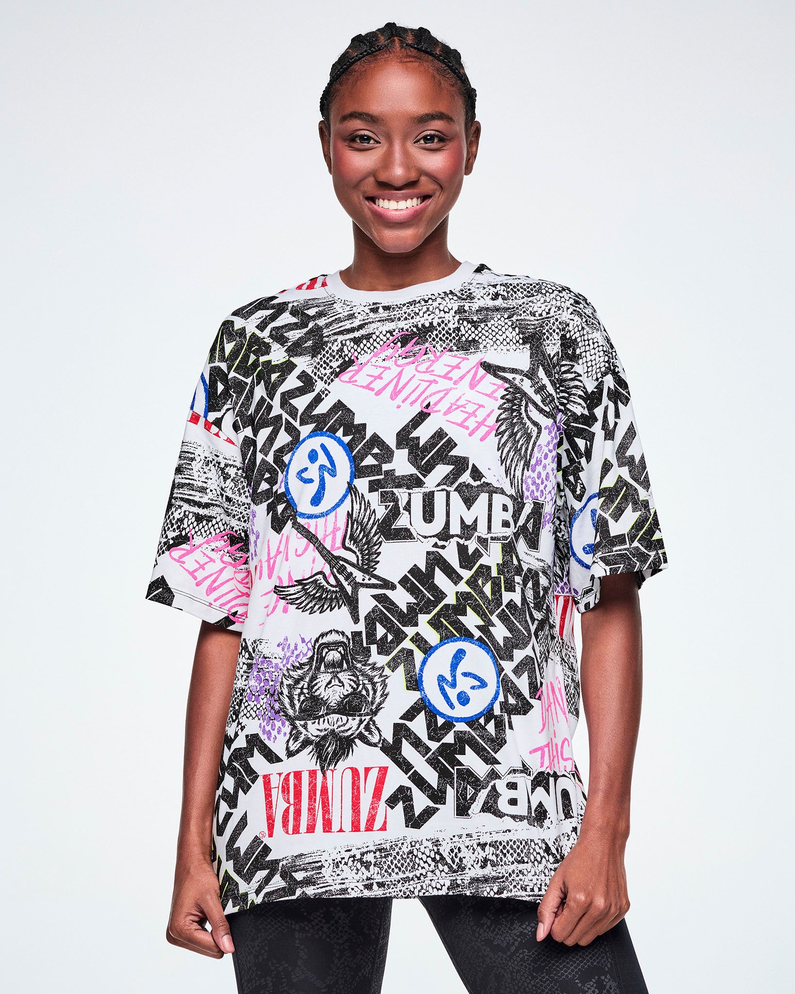 A smiling female model wearing a graphic tee with black, white, pink, and blue graffiti-style print and logos, such as blue zumbitos, a tiger wearing sunglasses, "Headliner energy, "Dance this Way," and ZUMBA in various rock and metal text, paired with black leggings.