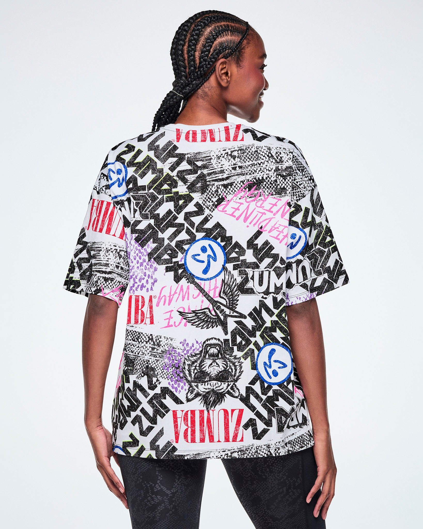 Back view of a female model wearing a graphic tee with black, white, pink, and blue graffiti-style print and logos, such as blue zumbitos, a tiger wearing sunglasses, "Headliner energy, "Dance this Way," and ZUMBA in various rock and metal text, paired with black leggings.