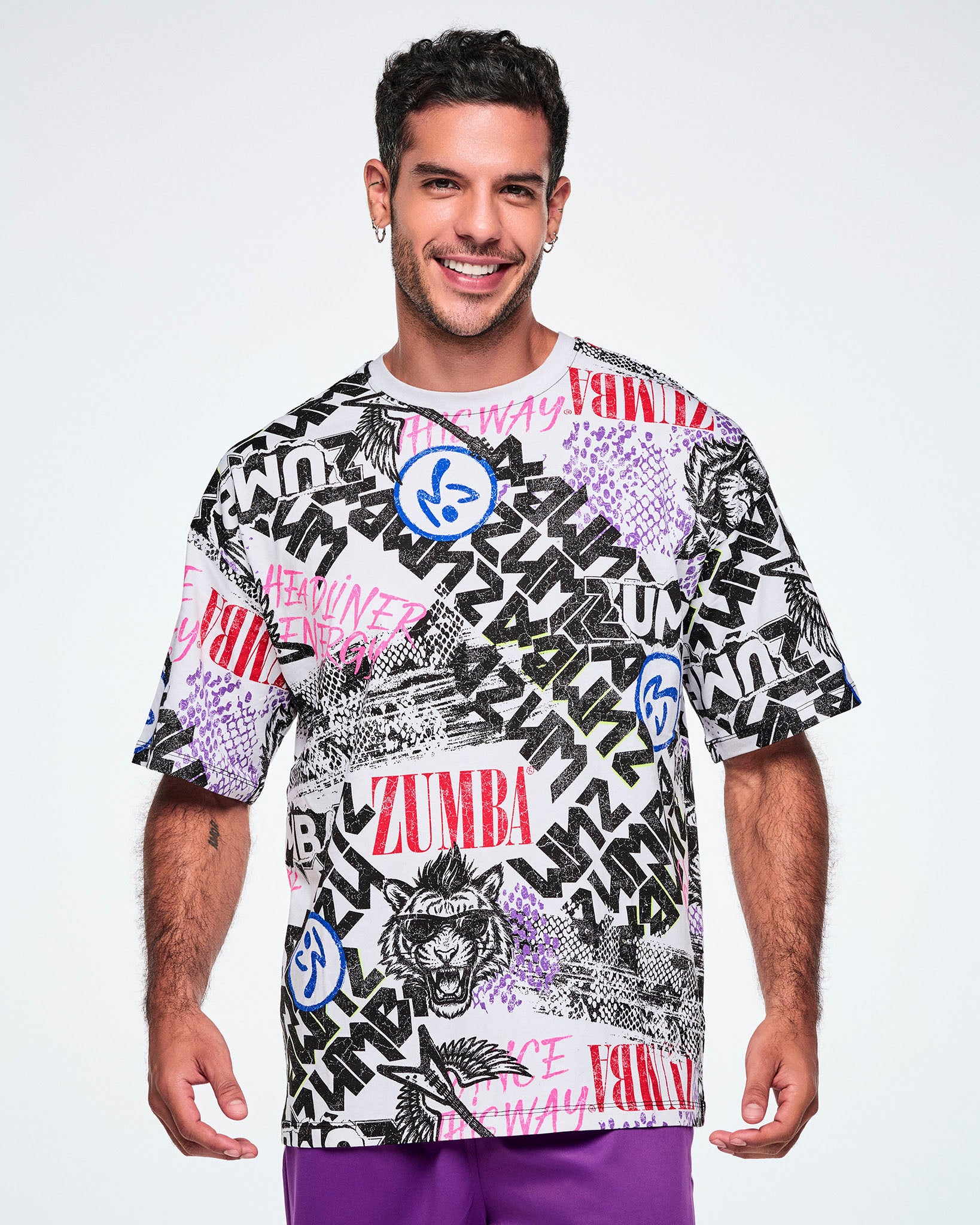 A smiling male model wearing a graphic tee with black, white, pink, and blue graffiti-style print and logos, such as blue zumbitos, a tiger wearing sunglasses, "Headliner energy, "Dance this Way," and ZUMBA in various rock and metal text, paired with purple pants.