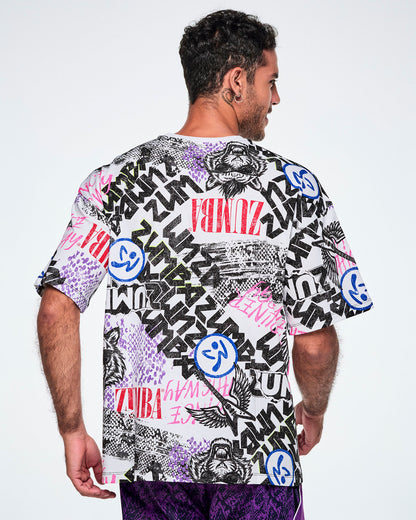 Back view of a male model wearing a graphic tee with black, white, pink, and blue graffiti-style print and logos, such as blue zumbitos, a tiger wearing sunglasses, "Headliner energy, "Dance this Way," and ZUMBA in various rock and metal text, paired with purple pants.