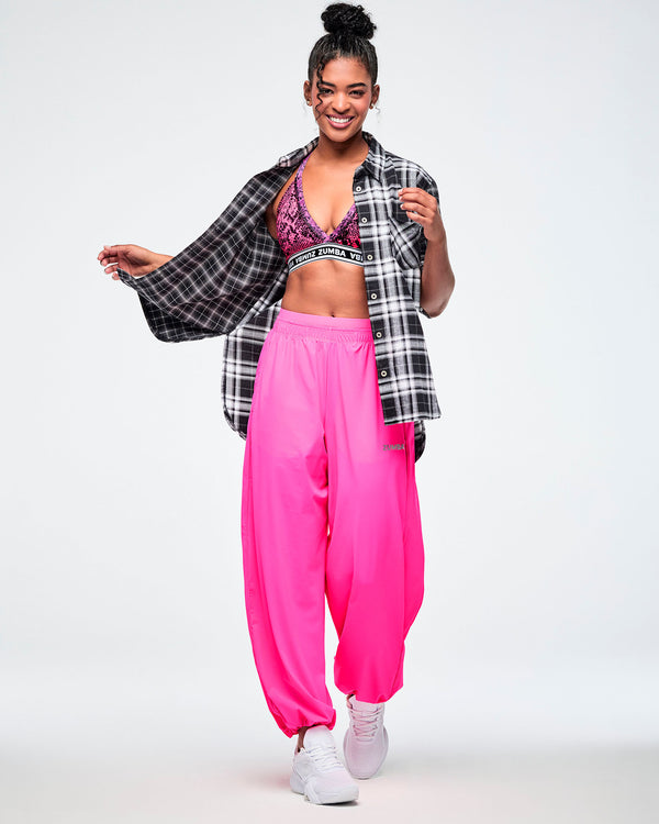 A female model wearing pink sweatpants, a black and white plaid button-down shirt, and a pink and black patterned sports bra, posing against a white background.