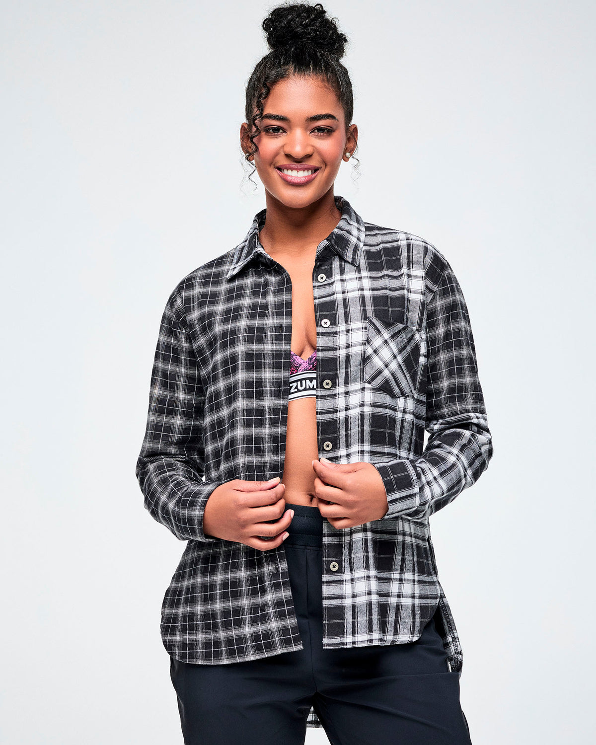 A model wears an open black-and-white plaid button-down shirt, unbuttoned to reveal a pink and black bra underneath, against a white background.