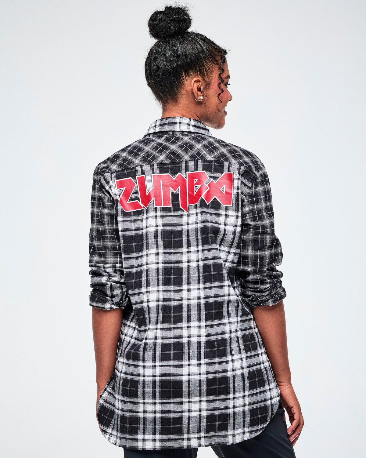 A female model shown from the back, wearing a black and white plaid button-down shirt with a red rock-like 'ZUMBA' text across the upper back, set against a white background. 