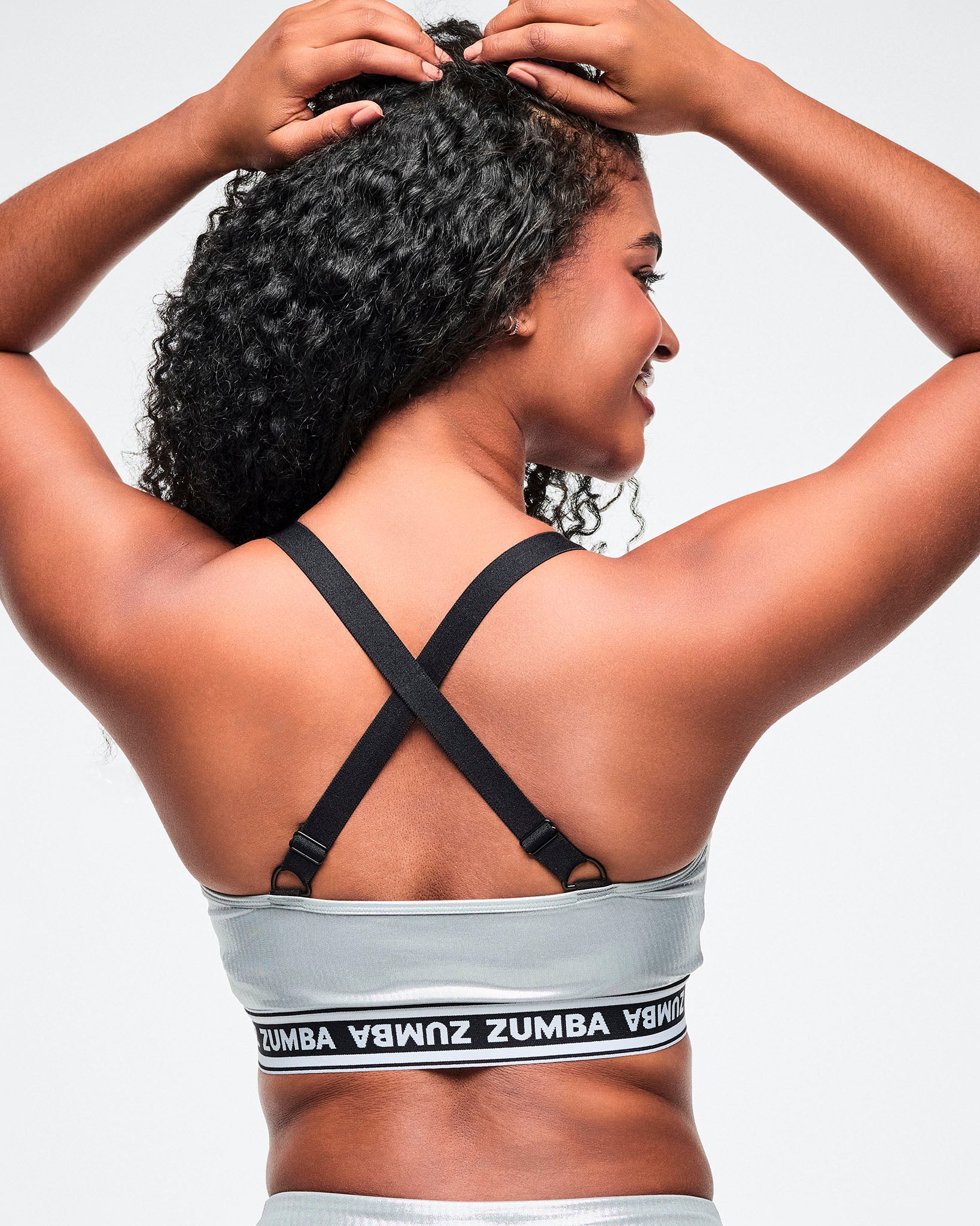 Back view of a female model with hands on her head, wearing a silver Zumba V‑neck sports bra with black crisscross adjustable straps and a black‑and‑white elastic band featuring repeating “ZUMBA” text.