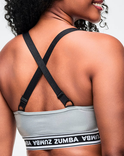 Back view of a female model wearing a silver Zumba V‑neck sports bra with black crisscross adjustable straps and a black‑and‑white elastic band featuring repeating “ZUMBA” text.