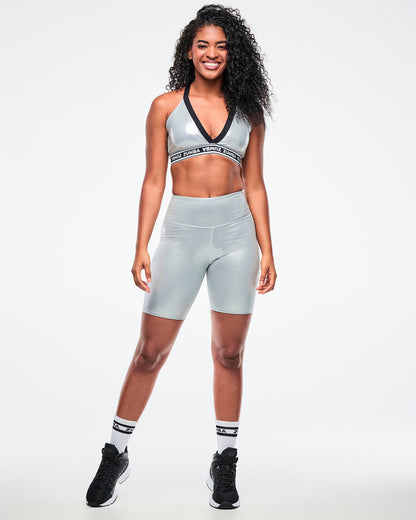 Female model wearing a silver Zumba V-neck sports bra with black trim and a black-and-white elastic band with repeating “ZUMBA” text, paired with silver biker shorts and black-and-white socks and shoes.