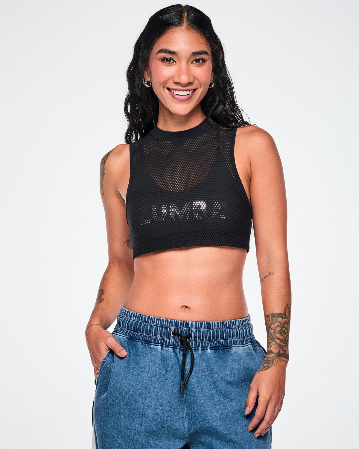 Front view of a female model wearing the black double-layer mesh Dale-Z crop bra, showing the silver "ZUMBA" logo on the inner layer visible through the mesh, paired with denim pants.