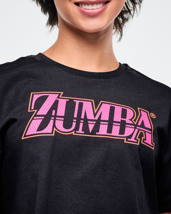 Close-up view of the female model in a black Zumba T-shirt with pink “ZUMBA” across the chest in a mesh-like pattern.