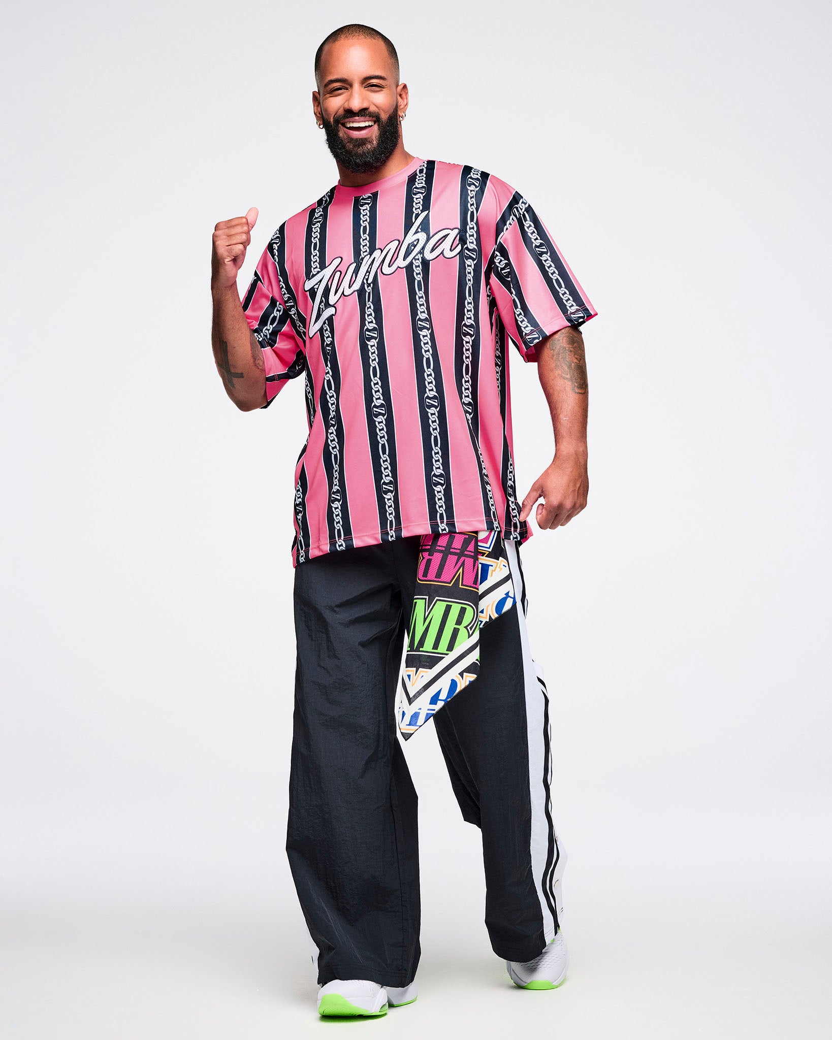 Male model wearing a pink-and-black striped Zumba top with white cursive “Zumba,” black-and-white striped pants, green-and-white shoes, and a bandana tucked into his pants.