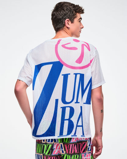 Back view of a male model in a white mesh top with large blue “ZUMBA” lettering and a pink Zumbito on the shoulder, paired with colorful pants against a white background.