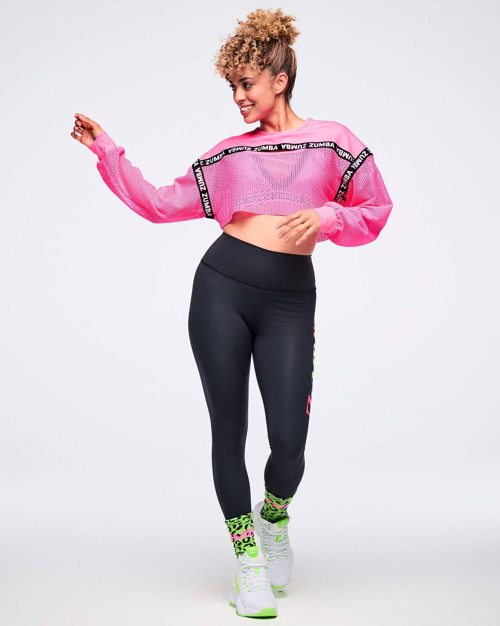 Female model dancing with swinging arms, wearing a pink cropped top, black leggings, green leopard-print socks, and green-and-white shoes.