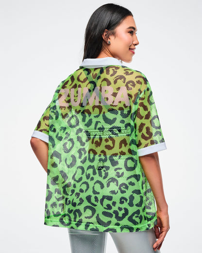 Back view of a green leopard‑print mesh polo with faint pink “Zumba” lettering across the back, paired with silver biker shorts.