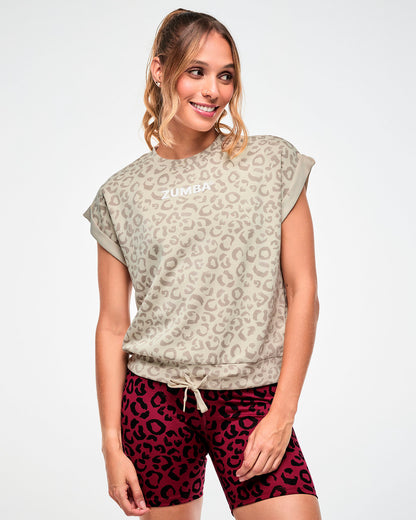Female model in a beige sleeveless sweatshirt with an all-over light brown leopard print, white 'ZUMBA' text on the chest, and a drawcord bottom band, paired with dark red leopard print biker shorts.