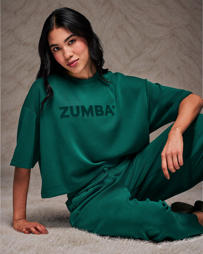 Female model reclining on a shaggy cream rug in the teal green drop shoulder crop top and matching pants. Taupe faux fur backdrop.