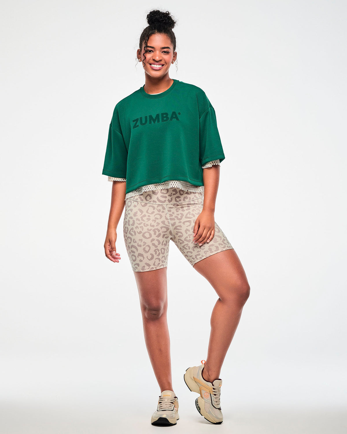 Full view of a female model in the teal green drop shoulder crop top layered over a beige mesh top, paired with beige leopard print biker shorts and beige shoes.