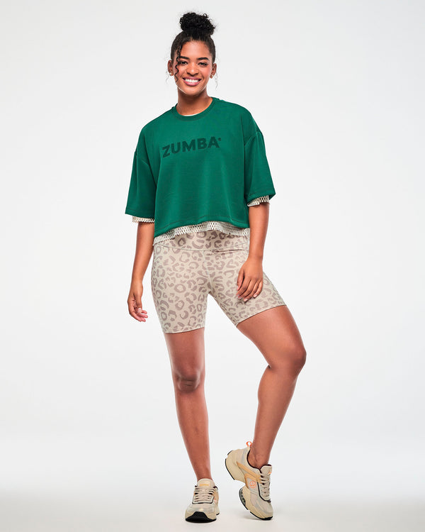 Full view of a female model in the teal green drop shoulder crop top layered over a beige mesh top, paired with beige leopard print biker shorts and beige shoes.