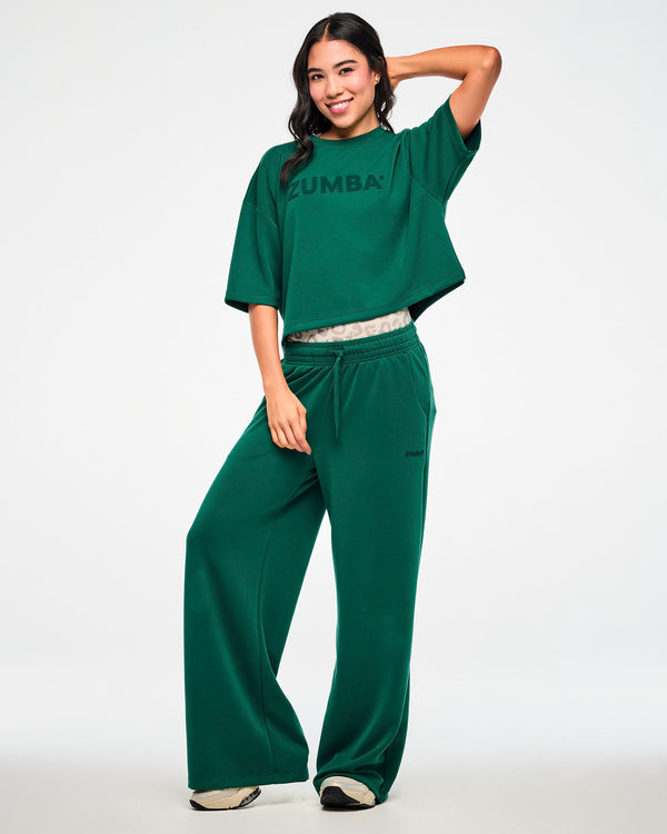 Full view of a female model with one hand behind her head, wearing the teal green crop top with matching teal high-waisted straight-leg pants and a beige leopard print waistband peeking out, paired with beige shoes.