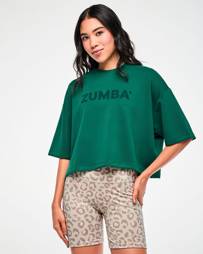 Female model in a teal green oversized drop shoulder crop top with large tonal 'ZUMBA' text across the chest, paired with beige leopard print biker shorts.