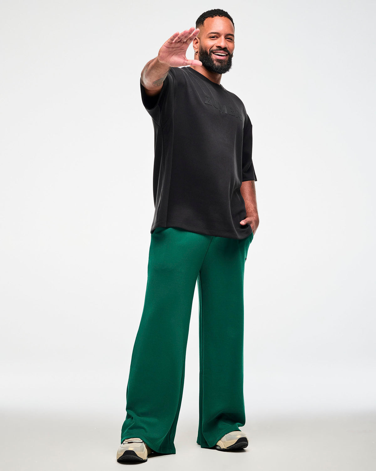 Full view of a male model reaching toward the camera in a black oversized drop shoulder top with tonal 'ZUMBA' text, paired with teal green wide-leg pants and beige shoes.